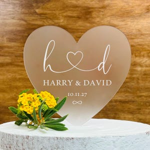 May include: A heart-shaped rose gold acrylic cake topper featuring white printed text that reads h heart d, HARRY & DAVID, 10.11.27, and an infinity symbol. A cluster of small yellow flowers is positioned at the base of the heart-shaped topper.