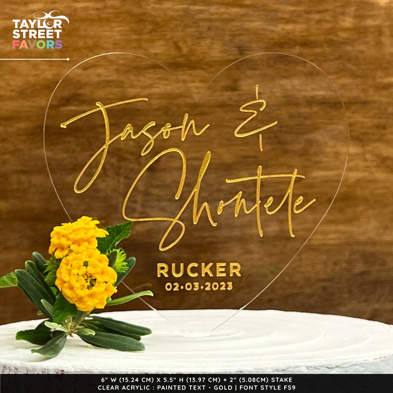 May include: A clear acrylic heart-shaped wedding cake topper with gold painted text. The names "Jason & Shontete" are displayed, along with "Rucker" and the date "02.03.2023". The topper is 6 inches (15.24 cm) wide and 5.5 inches (13.97 cm) high.