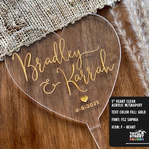 May include: A clear acrylic heart-shaped wedding cake topper with gold lettering that reads "Bradley & Karrah" and the date "4-8-2023". The topper is 5 inches in size and has a snap-off base.
