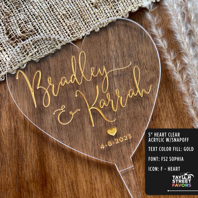 May include: A clear acrylic heart-shaped wedding cake topper with gold lettering that reads "Bradley & Karrah" and the date "4-8-2023". The topper is 5 inches in size and has a snap-off base.
