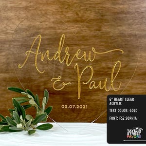 May include: A clear acrylic heart-shaped wedding cake topper with gold lettering that reads "Andrew & Paul" and the date "03.07.2021". The topper is 6 inches in size and features the text color gold and the font FS2 Sophia. A sprig of greenery is in the foreground.