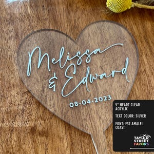 May include: A clear acrylic heart-shaped wedding cake topper with the names "Melissa & Edward" and the date "08-04-2023" in silver script. The topper is 5 inches in size and has a wooden handle.
