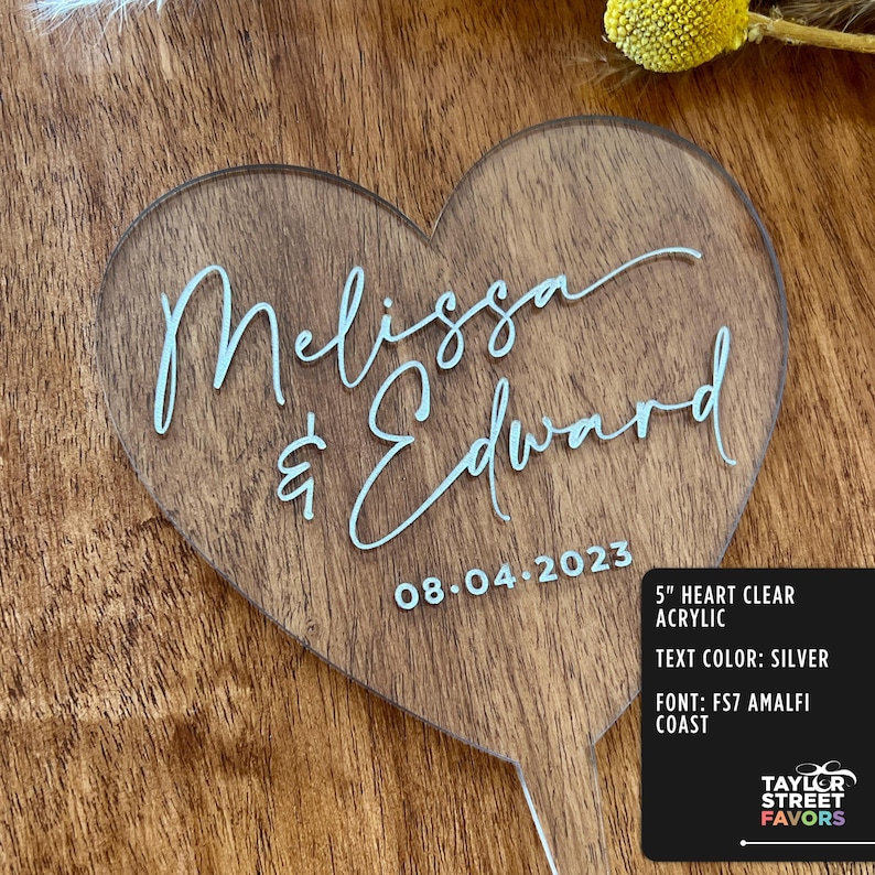 May include: A clear acrylic heart-shaped wedding cake topper with the names "Melissa & Edward" and the date "08-04-2023" in silver script. The topper is 5 inches in size and has a wooden handle.