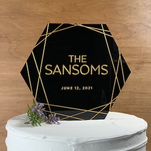 May include: Black hexagonal cake topper with gold geometric lines and the text "THE SANSOMS" and "JUNE 12, 2021".