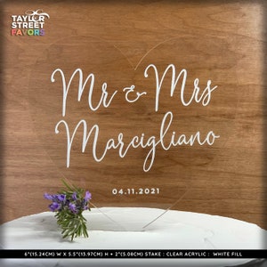 May include: Clear acrylic heart-shaped cake topper with the text "Mr & Mrs Marcigliano" and the date "04.11.2021". The topper is 6 inches (15.24cm) wide, 5.5 inches (13.97cm) high, and has a 2-inch (5.08cm) stake. A small bouquet of purple flowers is placed on the cake.