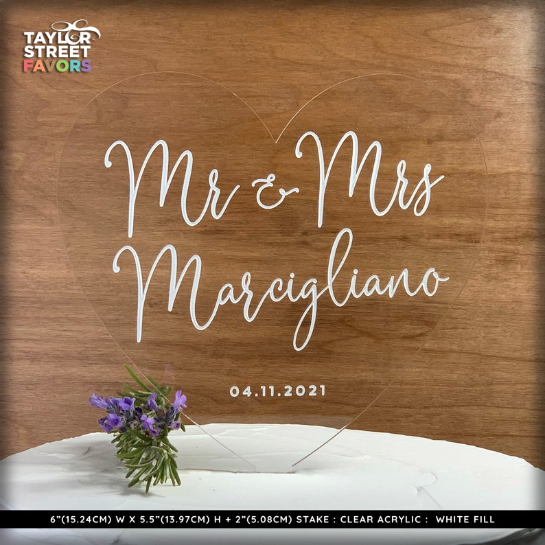 May include: Clear acrylic heart-shaped cake topper with the text "Mr & Mrs Marcigliano" and the date "04.11.2021". The topper is 6 inches (15.24cm) wide, 5.5 inches (13.97cm) high, and has a 2-inch (5.08cm) stake. A small bouquet of purple flowers is placed on the cake.