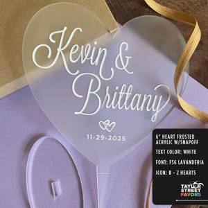 May include: A frosted acrylic heart-shaped cake topper with the names "Kevin & Brittany" in white script, along with the date "11.29.2025" and two heart icons. The topper is 6 inches in size and has a snap-off feature.