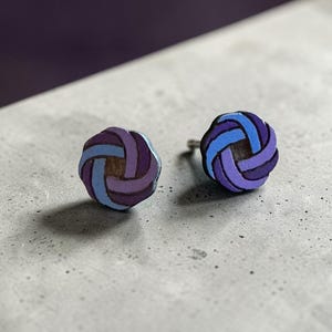 Op de afbeelding: Een paar ronde oorstekers. Elke oorbel heeft een geweven ontwerp in tinten paars, blauw en bruin. De oorbellen staan tegen een neutrale achtergrond, waardoor hun kleurrijke details worden benadrukt.