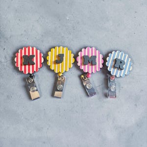 May include: Four circular badge reels with scalloped edges, each featuring vertical stripes in red, yellow, pink, or blue. Each reel displays a bold, black capital letter K, J, M, or R centred on the front, attached to a silver metal clip.