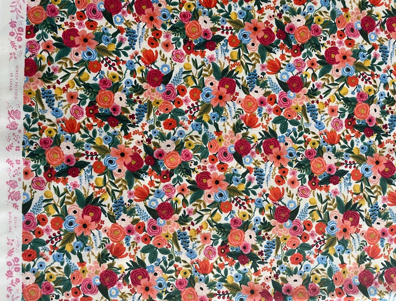 May include: A white fabric with a floral pattern featuring red, pink, blue, and green flowers. The fabric is covered in a variety of flowers and leaves.
