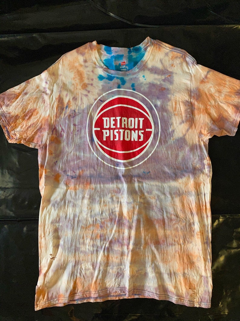 May include: A tie-dye t-shirt with a white basketball logo that says "DETROIT PISTONS" in red letters.