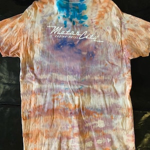 May include: A tie-dye t-shirt with an orange, blue, and purple color scheme. The shirt has the text "Motor City Casino-Hotel" printed on the back.