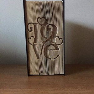 May include: A book art sculpture of the word "LOVE" with hearts. The book is folded into a three-dimensional shape and is displayed on a wooden surface.