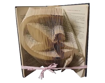 Può includere: Un libro con le pagine piegate in un design 3D di un uccello. Il libro è aperto e l'uccello è in piedi sulle pagine. Il libro è legato con un nastro rosa.