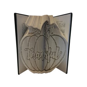 May include: A folded book art sculpture featuring a pumpkin shape with the word Thankful displayed across the centre. The pages are precisely creased to form the design, with the black book covers visible on the left and right sides of the sculpture.
