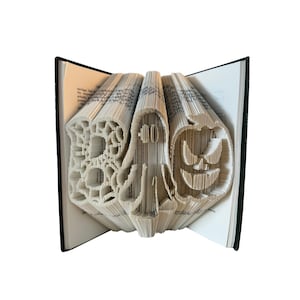 Peut inclure: Une sculpture en livre plié représentant le mot BOO, une figure de fantôme centrale et un motif de citrouille lanterne. Les pages sont précisément pliées et marquées pour créer ces formes tridimensionnelles dans la structure du livre ouvert.