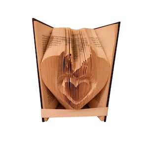 Peut inclure: Une sculpture de livre plié dont les pages sont minutieusement marquées pour former une forme de cœur dans une autre forme de cœur. Le livre a des couvertures marron foncé et une bande de papier clair enroulée à la base pour maintenir la structure.