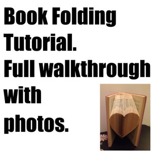 May include: A book folded into the shape of a heart. The book is open and the pages are folded to create a three-dimensional heart shape. The book is on a table with a pink surface. The text "Book Folding Tutorial. Full walkthrough with photos." is visible above the book.
