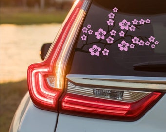 May include: A vinyl decal applied to a vehicle rear window featuring a black tree branch silhouette adorned with numerous light pink cherry blossom flowers. The design spans horizontally across the glass, showcasing detailed five-petaled blooms.