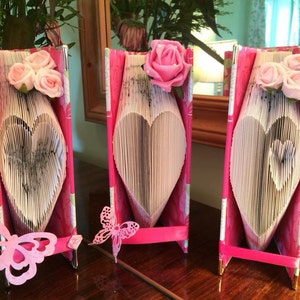 Puede incluir: Tres libros en forma de corazón rosa y blanco con flores rosas en la parte superior. Cada libro está sobre un soporte rosa con una decoración de mariposa.
