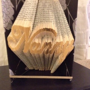 May include: A book art sculpture featuring the name "Natalie" folded into the pages. The sculpture is displayed on a black base with a silver and black patterned background.