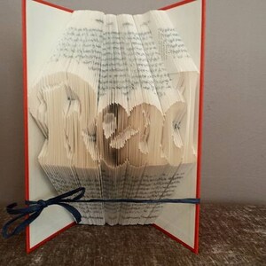 May include: A book with the word "Read" folded into the pages. The book is open and standing upright on a table. The book has a red cover and a blue ribbon.