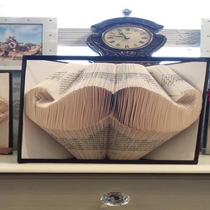 Puede incluir: Una escultura de libro con páginas dobladas para crear la forma de un bigote. El libro se exhibe en un marco negro, colocado frente a un reloj analógico oscuro con números romanos y flanqueado por dos marcos blancos con fotografías.