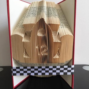 May include: A book art sculpture of a racing car made from the pages of a book. The book is open and the pages are folded into the shape of a racing car. The book is bound in red and has a black and white chequered ribbon around the bottom.