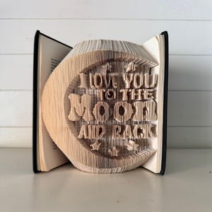 May include: A folded book art sculpture featuring the text I LOVE YOU TO THE MOON AND BACK. The pages are precisely creased and folded to form the letters and a crescent moon shape, with small star designs positioned above and below the text.