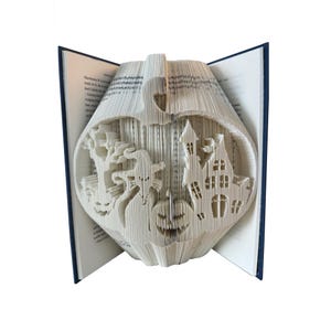 May include: A folded book art sculpture featuring a large pumpkin shape containing intricate cutouts of a haunted house, a witch, a tree, and a smaller jack-o'-lantern. The pages are precisely folded to create these detailed silhouettes.