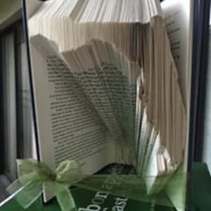 May include: A book art sculpture shaped like a state, possibly Florida, with pages folded into a three-dimensional design. The book is open and the pages are folded to create the shape of the state. The book is sitting on a green book with the title "bon appétit fast, fresh, easy" visible on the spine.