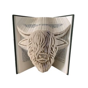 May include: A book sculpture featuring a Highland cow design carved into the pages. The open book reveals the cow's head, with detailed cuts forming its features. The book has a dark green cover and the pages are a light cream colour.