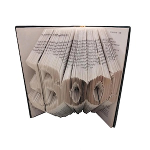May include: A book sculpture featuring pages folded to form the word BOO! in large, three-dimensional letters. The book is open with a dark cover, and the white paper pages are precisely creased to create the text, which is clearly visible.