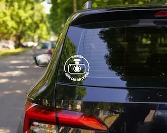 May include: A white warning sticker on a black car's rear window. The sticker reads "Warning Dash Cam Recording" with a camera icon inside a circle.
