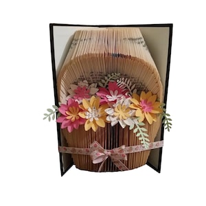 May include: A book art sculpture of a flower arrangement in a basket. The basket is made from the pages of a book that has been folded and shaped. The flowers are made from paper and are in shades of pink, yellow, and green.