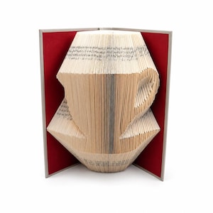 Puede incluir: Una escultura de libro con páginas dobladas que forman la silueta de una taza de café. El libro tiene una cubierta dura de color gris claro con un forro interior rojo intenso. Las páginas de papel crema muestran texto negro impreso.
