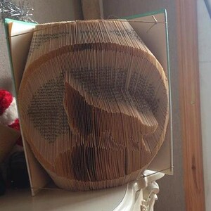 May include: A book folded into the shape of a bird, with the pages forming the bird's body and wings. The book is made of brown paper and has a green cover.