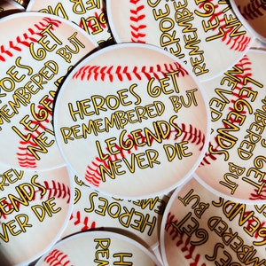 May include: Circular stickers with a baseball design. The stickers feature a baseball graphic with red stitching and the text "Heroes Get Remembered But Legends Never Die" in a yellow font with a black outline.