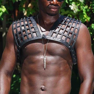 May include: A black leather harness with silver spikes is worn over the chest. The model is wearing a pair of iridescent blue and purple leggings. The outfit is completed with black fingerless gloves and sunglasses. A silver pendant hangs from a necklace.