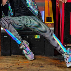 May include: A person wearing a black shirt, a silver sequined jacket, and iridescent leggings with a black polka dot pattern. They are wearing a tall black top hat with a silver band. The person is sitting on a black bench in front of a red and black arcade game with the words "Super Shot" on it.