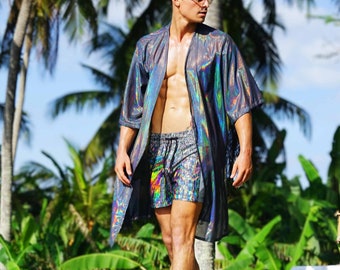 May include: A man wearing a iridescent silver and black kimono with a colorful patterned swim short. He is standing in front of palm trees.