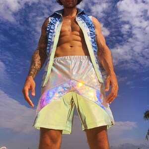 May include: A person wearing a light blue sleeveless shirt with ruffles and iridescent green and silver shorts. The person is standing in front of a blue sky with white clouds.