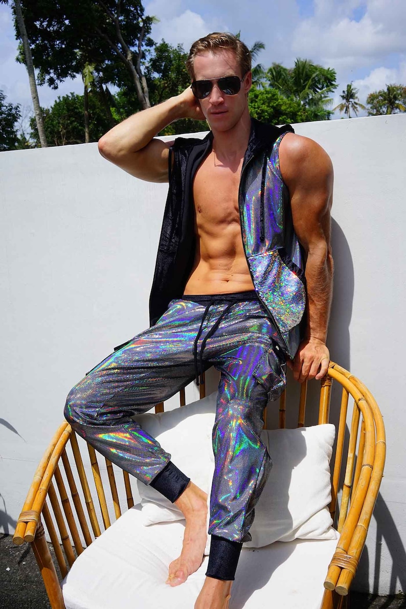 May include: A man wearing a black and iridescent sleeveless hoodie and iridescent pants. He is sitting on a bamboo chair with a white cushion.