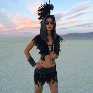 May include: A woman wearing a black feather headdress, a black leather hooded top with feather trim, a black leather skirt with feather trim, and sunglasses. She is standing in a desert landscape.
