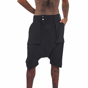 May include: Black shorts with a unique design. The shorts feature a high waistband with two buttons and two large pockets. The shorts have a loose, draped fit, and the fabric appears to be a lightweight material.