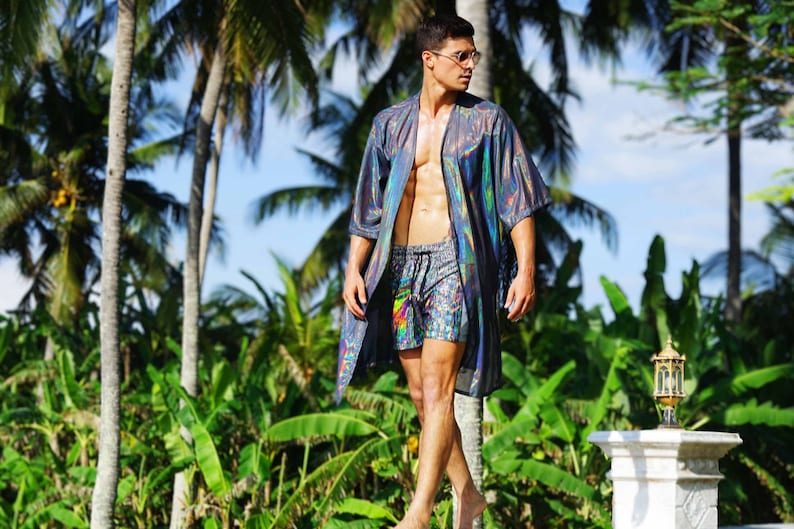 K&ouml;nnte beinhalten: Ein Mann tr&auml;gt einen regenbogenfarbenen silbernen und schwarzen Kimono mit einer bunten gemusterten Badehose. Er steht vor Palmen.