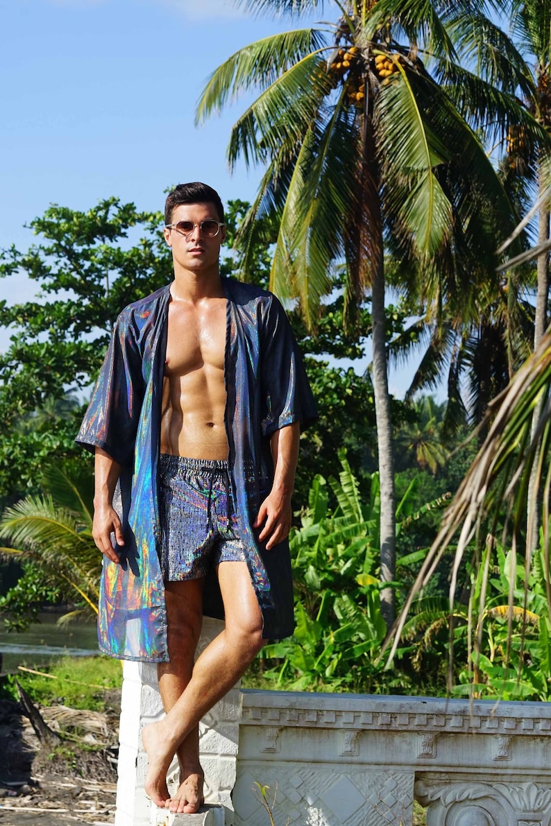 K&ouml;nnte beinhalten: Ein Mann tr&auml;gt einen regenbogenfarbenen blau-schwarzen Kimono mit einer passenden Badehose. Er steht vor einem Palmenbaum und einer wei&szlig;en Steinmauer.