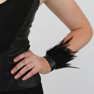 May include: A black leather bracelet with a black feather trim. The bracelet is worn on the wrist of a person.