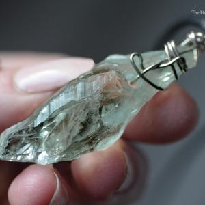 May include: A pale green crystal pendant with a silver wire wrap. The crystal is irregularly shaped and has a textured surface. The pendant hangs from a black cord. The Healing Sea Wolf is in the upper right corner.