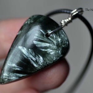 May include: A teardrop-shaped pendant made of dark green seraphinite stone with silver-colored fibrous inclusions. The stone is suspended from a black cord by a silver-toned wire wrap. The text The Healing Sea Wolf is visible in the upper right.